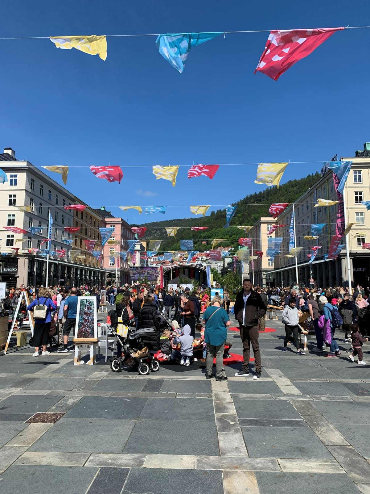 A crowded city street under strung flags, stalls along both sides and a mountain rising behind the rooftops